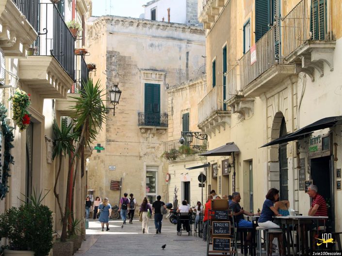Une rue étroite dans une vieille ville européenne, à Lecce en Italie, avec des terrasses de café des deux côtés. Plusieurs personnes se promènent et dînent, tandis qu'un oiseau se tient sur le trottoir. Des balcons et des plantes bordent les bâtiments.