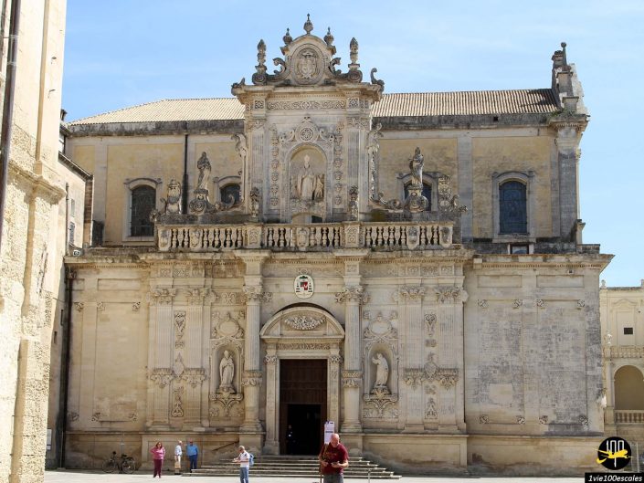 Façade de bâtiment historique de style baroque, à Lecce en Italie, avec des sculptures et des gravures ornées, avec une entrée centrale. Plusieurs personnes sont visibles devant la structure, sur un ciel bleu clair en arrière-plan.