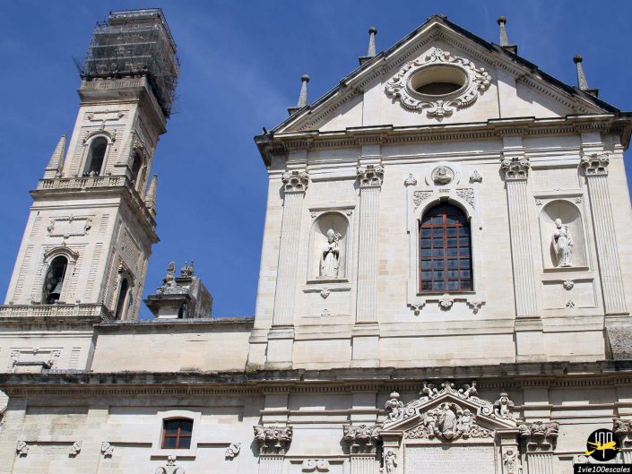 Bâtiment en pierre avec des détails architecturaux richement ornés, notamment des statues et des fenêtres cintrées, à Lecce en Italie. La tour adjacente est en construction, recouverte d'échafaudages. Ciel bleu en arrière-plan.