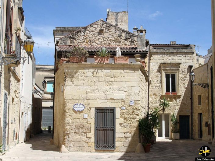 Une rue étroite en pierre avec des bâtiments anciens dotés de balcons et de plantes en pot, à Lecce en Italie. Les bâtiments ont des pierres beiges patinées et quelques petites fenêtres. Le ciel est clair et bleu.