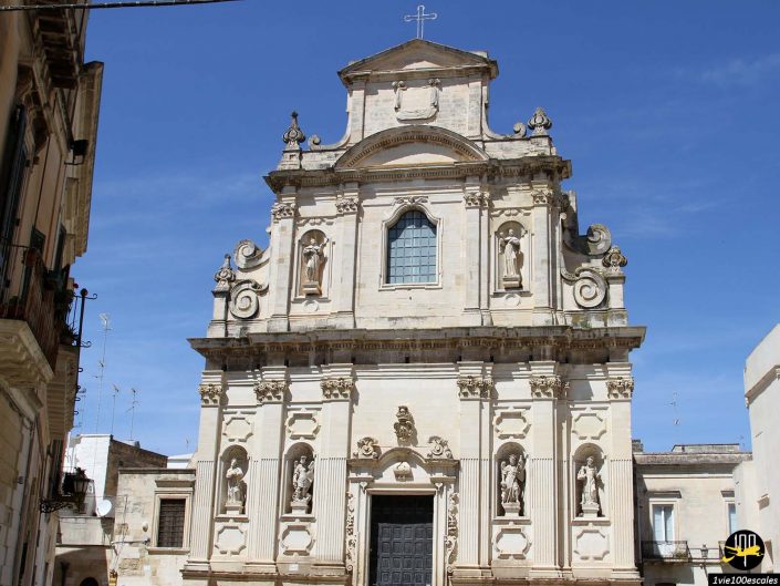 Une église en pierre beige avec des sculptures et des colonnes ornées, avec une entrée centrale voûtée, des statues décoratives dans des niches et une croix au sommet, sur un ciel bleu clair à Lecce en Italie.