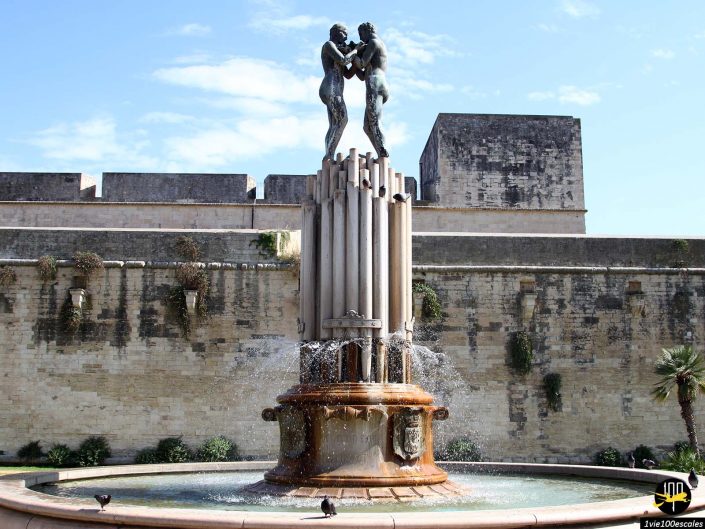 Une grande fontaine avec des statues de deux personnages près du sommet se dresse dans un espace ouvert, adossée à un vieux mur de pierre à Lecce en Italie. La fontaine comporte plusieurs jets d'eau et des pigeons rassemblés à sa base.