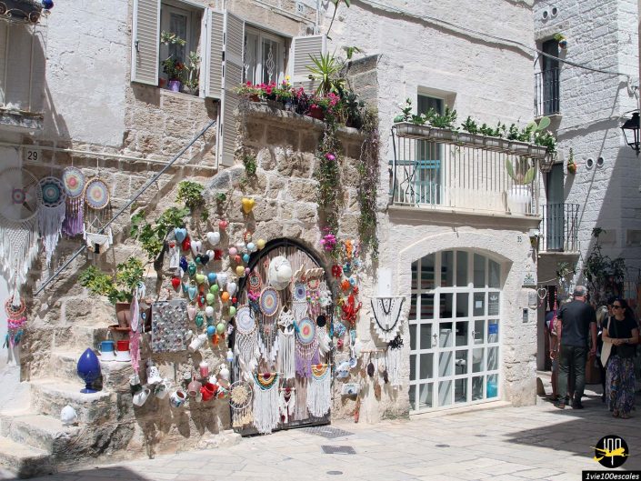 Un bâtiment en pierre décoré d'attrape-rêves et de bibelots colorés est présenté. Plusieurs plantes ornent le balcon et l'entrée, à Polignano a Mare en Italie. Deux personnes marchent sur le côté droit du bâtiment.
