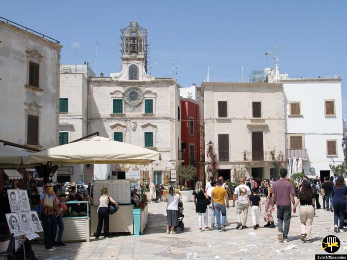 Un marché en plein air animé sur une place de ville européenne, à Polignano a Mare en Italie, avec des bâtiments historiques, une tour de l'horloge et divers étals de marché avec des gens marchant et parcourant sous un ciel bleu clair.