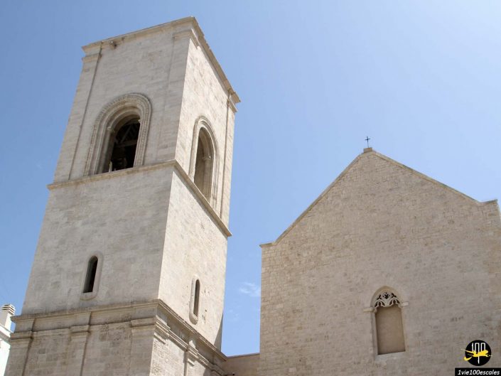 Un haut clocher en pierre et une grande église en pierre, chacune avec des fenêtres rectangulaires et cintrées, se dressent majestueusement sur un ciel bleu clair à Polignano a Mare en Italie.