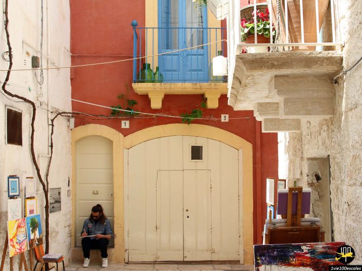 Une personne est assise sur un banc contre un mur rougeâtre avec un balcon bleu au-dessus. De nombreuses peintures sont exposées le long de la ruelle étroite, capturant le charme de Polignano a Mare en Italie.