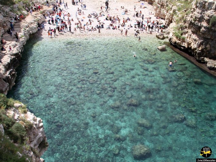 Une plage bondée au bord d'un bassin rocheux aux eaux turquoise et claires, qui rappelle Polignano a Mare en Italie. Les nageurs profitent de l'eau rafraîchissante tandis que de nombreuses personnes sont assises et debout sur la rive sablonneuse et les rochers environnants.
