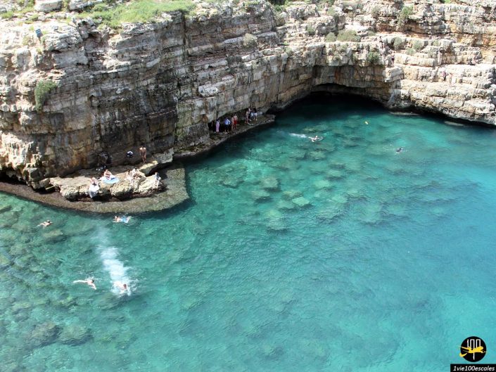 Une baie aux eaux claires émeraude entourée de falaises rocheuses avec des gens nageant et assis sur les rochers, à Polignano a Mare en Italie. Certains individus sont rassemblés dans une petite zone semblable à une grotte dans la falaise.