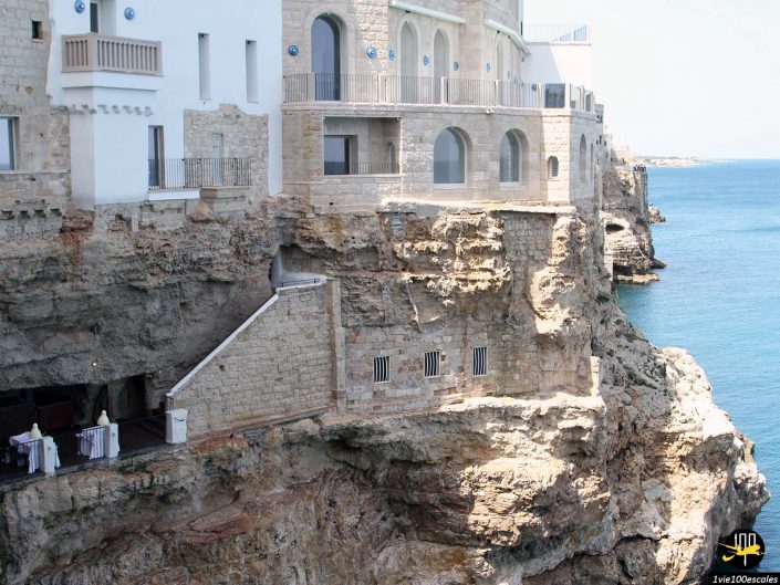 Un bâtiment blanc et en pierre avec des fenêtres cintrées se dresse au sommet d'une falaise rocheuse surplombant une mer bleue et calme, à Polignano a Mare en Italie. Des escaliers descendent du bâtiment jusqu'au niveau inférieur de la falaise.