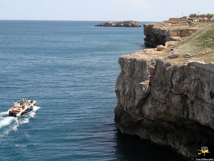 Un petit bateau voyage sur une eau bleu clair près d'une falaise rocheuse, avec des gens assis et marchant au bord de la falaise sous un ciel clair à Polignano a Mare en Italie.