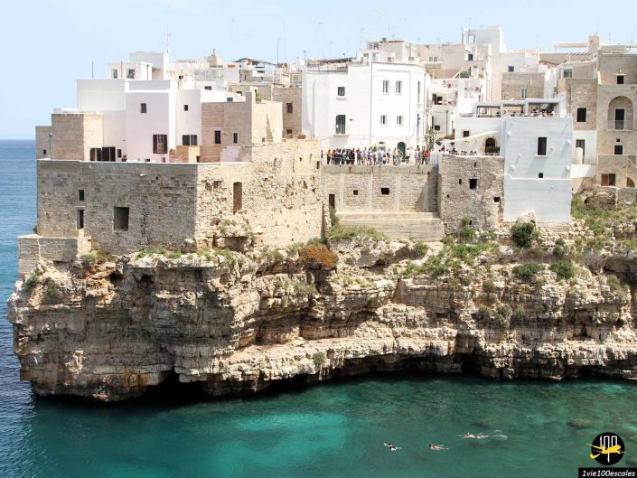 Une ville côtière aux bâtiments blancs et aux structures en pierre perchée sur une falaise rocheuse surplombant une eau bleu clair ; on voit quelques personnes nager près de la falaise, qui rappelle à Polignano a Mare en Italie.