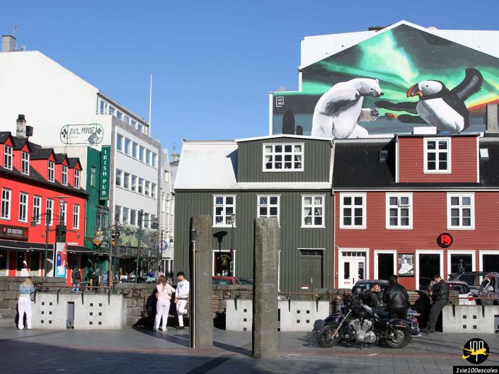 Une scène de rue à Reykjavik en Islande avec des bâtiments colorés, des gens debout et assis, deux motos au premier plan et une fresque représentant un ours polaire et un macareux sur l'un des bâtiments.
