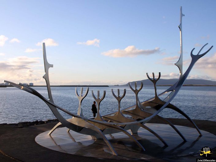 Une grande sculpture métallique ressemblant à un bateau viking stylisé avec plusieurs sections pointant vers le haut est située au bord d'un front de mer avec des montagnes en arrière-plan, à Reykjavik sur l'île. Une personne se tient à proximité.