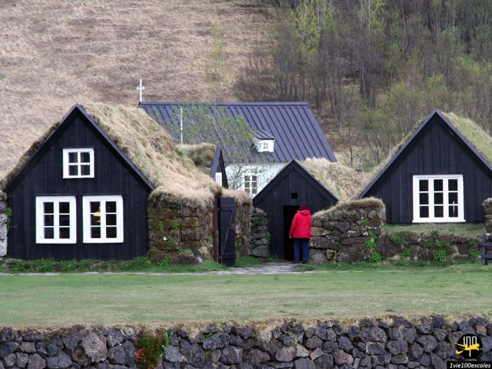 Une personne en manteau rouge se tient devant des maisons en bois noir au toit de tourbe et des murs en pierre dans une zone herbeuse à Reykjavik en Islande.