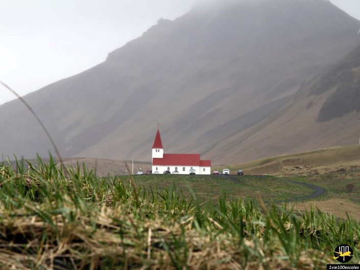 Une petite église blanche au toit rouge se trouve sur une colline avec un terrain montagneux et du brouillard en arrière-plan, typique d'en Islande. L'herbe verte et les voitures garées sont visibles au premier plan.