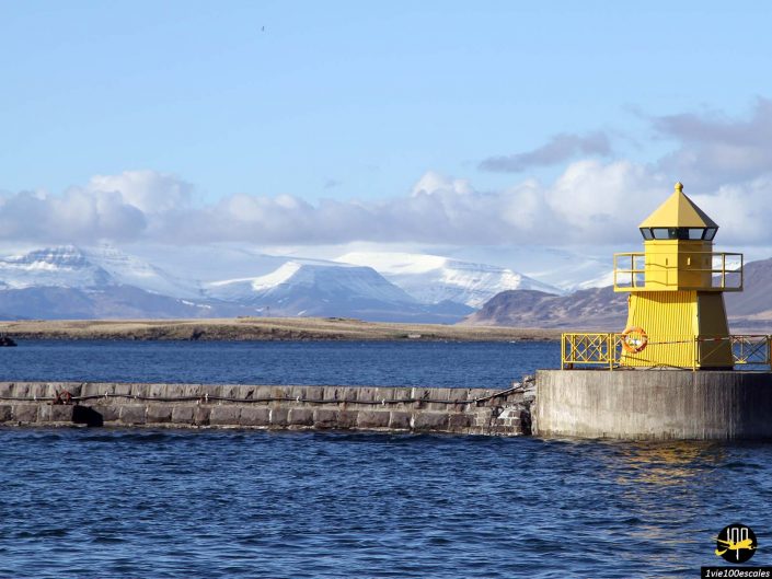 Un phare jaune se dresse sur une jetée en béton dans une mer calme avec des montagnes enneigées et un ciel partiellement nuageux en arrière-plan, à Reykjavik en Islande.
