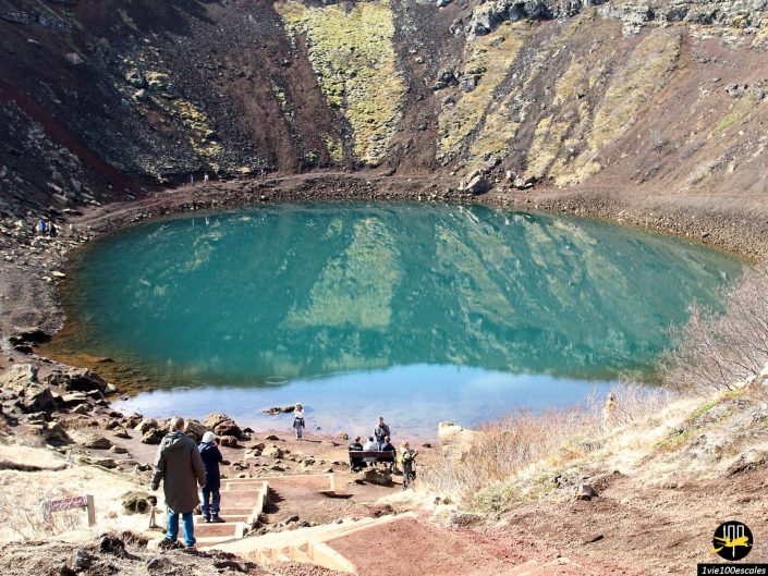 Les gens descendent un escalier en bois jusqu'au bord d'un lac de cratère turquoise entouré de falaises rocheuses avec un peu de végétation, en Islande.