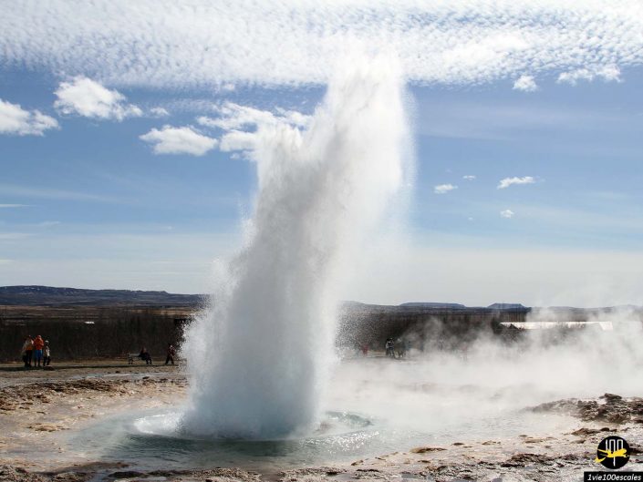 Un puissant geyser entre en éruption, envoyant une colonne d'eau et de vapeur vers le ciel dans le paysage aride d'en Islande sous un ciel partiellement nuageux.