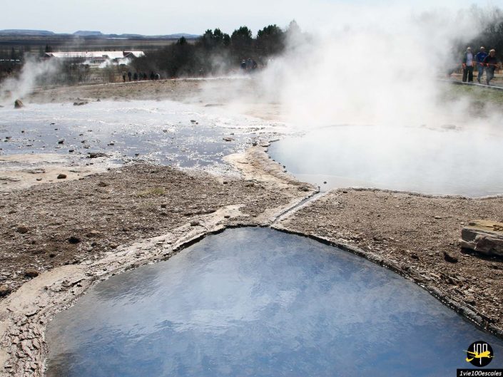 Sources chaudes géothermiques avec vapeur s'élevant de bassins bleus entourés d'un terrain rocheux à Islande. Les visiteurs sont visibles en arrière-plan près des arbres.