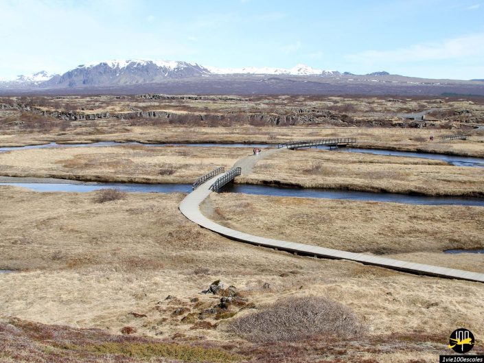 Une promenade en bois s'étend à travers un vaste paysage aride en Islande, avec des montagnes lointaines sous un ciel bleu clair. Une végétation clairsemée et des ruisseaux peu profonds sont visibles. On voit quelques personnes marcher sur la promenade.