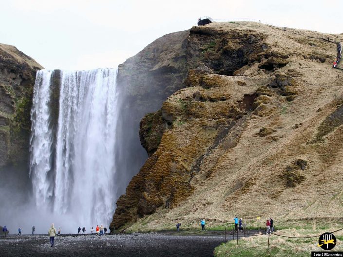 Une grande cascade tombe sur une falaise en Islande, avec des gens debout à sa base sur une zone rocheuse. Certaines personnes gravissent le flanc de la colline. Le paysage est aride avec une végétation clairsemée.