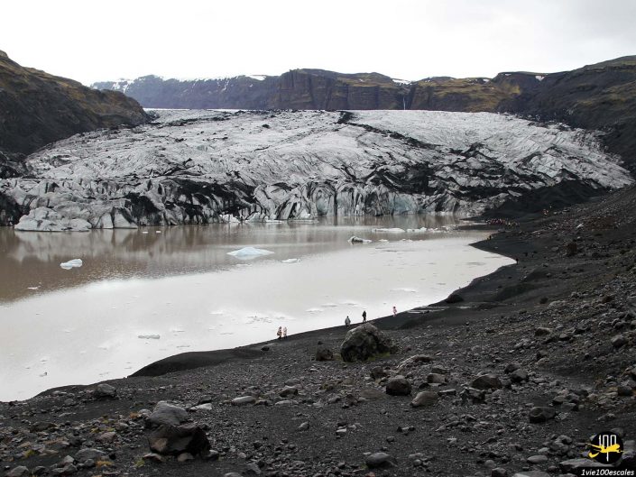 Vue d'un grand glacier descendant dans un lac partiellement gelé, entouré d'un terrain rocheux sombre, avec quelques petites figures visibles près du bord de l'eau en Islande.