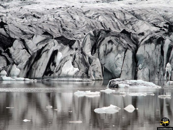 Un glacier avec des stries sombres et de gros morceaux de glace flottant dans une étendue d'eau, trouvé en Islande. La glace est un mélange de noir, de gris et de blanc qui se reflète sur la surface de l’eau.