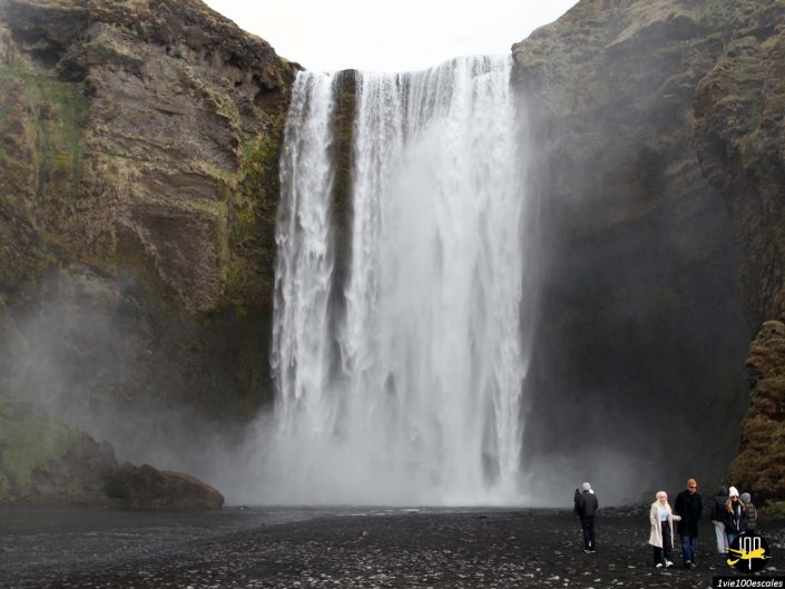Un groupe de personnes se tient près de la base d’une haute cascade tombant en cascade sur le flanc d’une falaise, entourée de brume et d’un terrain rocheux en Islande.