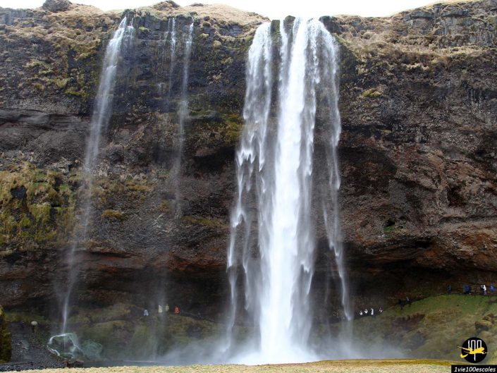 Une grande cascade tombe en cascade sur une falaise rocheuse avec des zones couvertes de mousse à Islande, et un groupe de personnes peut être vu près de la base.