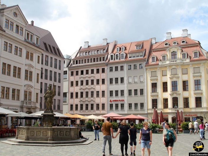 Une place pavée à Dresde en Allemagne avec des passants et une fontaine au centre, entourée de grands bâtiments historiques et de cafés en plein air.