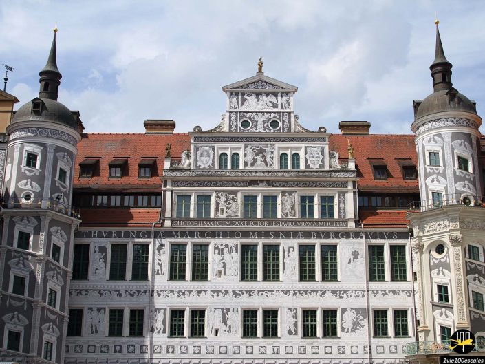 Un bâtiment historique avec des détails architecturaux complexes et des motifs en pierre blanche et grise, à Dresde en Allemagne, avec deux tours cylindriques et un toit de tuiles rouges.