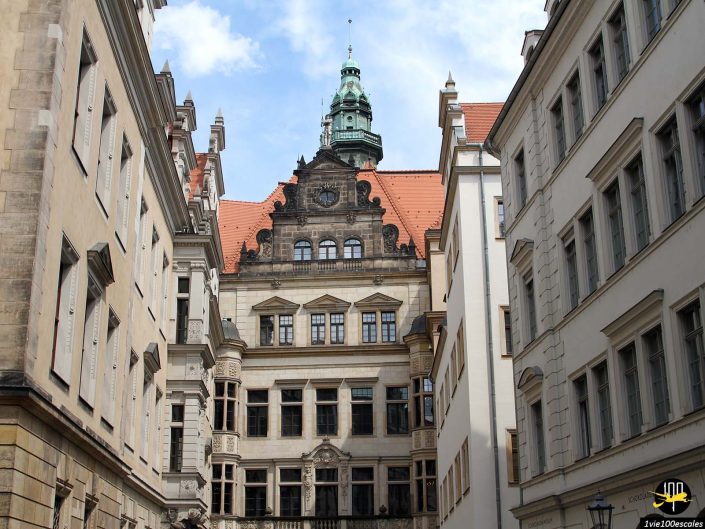 Un bâtiment historique à l'architecture richement ornée, doté d'une tour centrale, de fenêtres cintrées et d'un toit rouge abrupt, encadré par des bâtiments majestueux adjacents à Dresde en Allemagne.