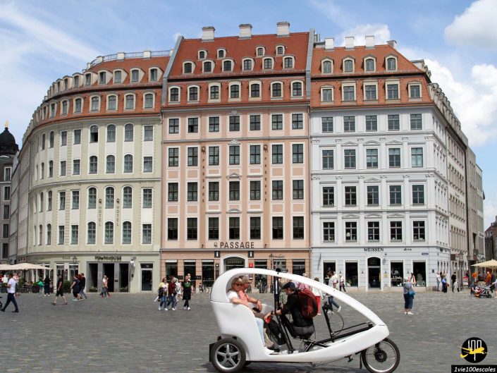 Une place animée de Dresde en Allemagne présente des bâtiments historiques à plusieurs étages et un taxi à pédales blanc moderne avec des passagers au premier plan.