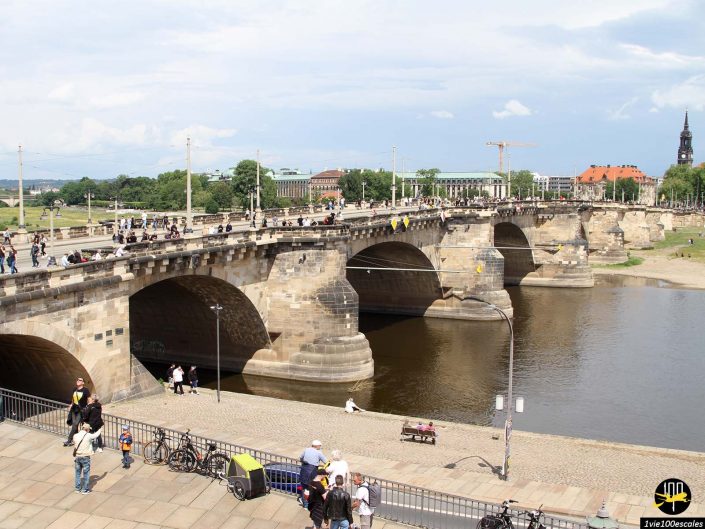 Un pont de pierre à arches multiples enjambe une rivière à Dresde en Allemagne. Les gens marchent le long du pont et de la promenade. Des bâtiments et des arbres sont visibles en arrière-plan sous un ciel partiellement nuageux.