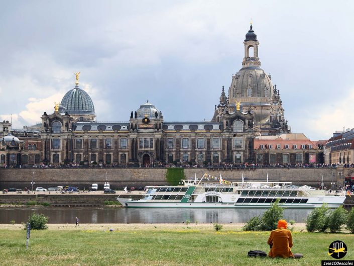 Une personne est assise sur l'herbe près de l'Elbe à Dresde en Allemagne, face à l'Académie des Beaux-Arts de Dresde et à la Frauenkirche, avec un ferry blanc et vert naviguant le long du fleuve.