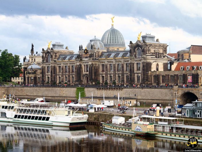 Un grand bâtiment historique avec un toit en forme de dôme se trouve le long d'une rivière à Dresde en Allemagne. Plusieurs bateaux sont amarrés au premier plan, avec des piétons et des cyclistes à proximité. Le ciel est partiellement nuageux.