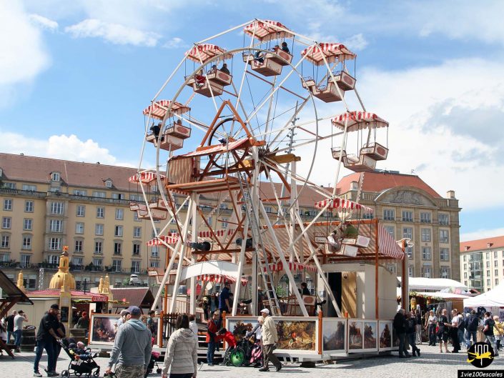 Les gens sont rassemblés autour d'une grande roue vintage sur un marché en plein air, avec des bâtiments historiques en arrière-plan, par une journée ensoleillée à Dresde en Allemagne.