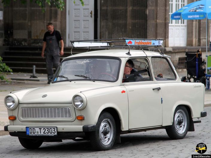 Une voiture Trabant blanche avec la plaque d'immatriculation "DD T 2303" est garée dans une rue pavée à Dresde en Allemagne. Une personne est visible à l’intérieur de la voiture, tandis qu’un homme en salopette se tient à l’arrière-plan.