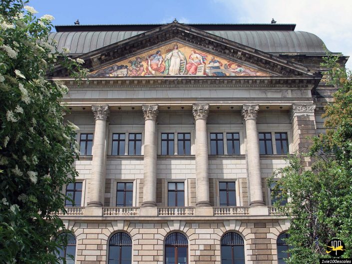 La vue de face d'un bâtiment classique orné de colonnes, de fenêtres cintrées et d'une frise colorée à Dresde en Allemagne. Les arbres flanquent le premier plan.