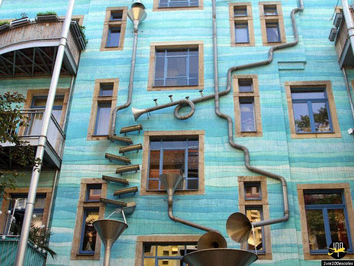 Un bâtiment turquoise vibrant à Dresde en Allemagne présente un système complexe de tuyaux et d'entonnoirs métalliques fixés à sa façade, probablement conçu comme un système unique de drainage des eaux de pluie.