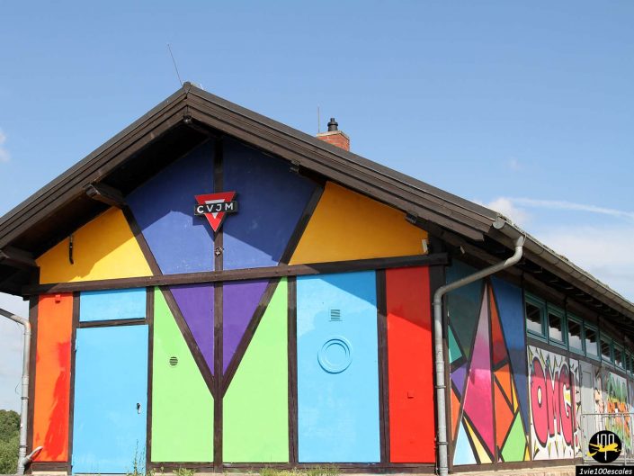 Un bâtiment coloré à Dresde en Allemagne avec des motifs géométriques et une enseigne CVJM sur la façade. La structure présente des panneaux bleus, orange, violets, rouges, jaunes et verts, avec des graffitis ornant la paroi latérale.